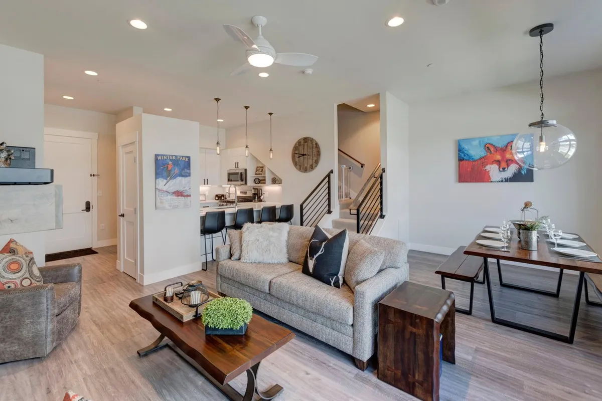 The image showcases an open-concept living area filled with natural light, featuring a plush sofa perfect for cozy movie nights, a stylish dining table ideal for family meals or entertaining friends, bar stools at a modern kitchen island, and tasteful mountain-inspired artwork on the wall. The inviting atmosphere is both warm and stylish, creating a welcoming space for groups to relax, dine, and make memories together.