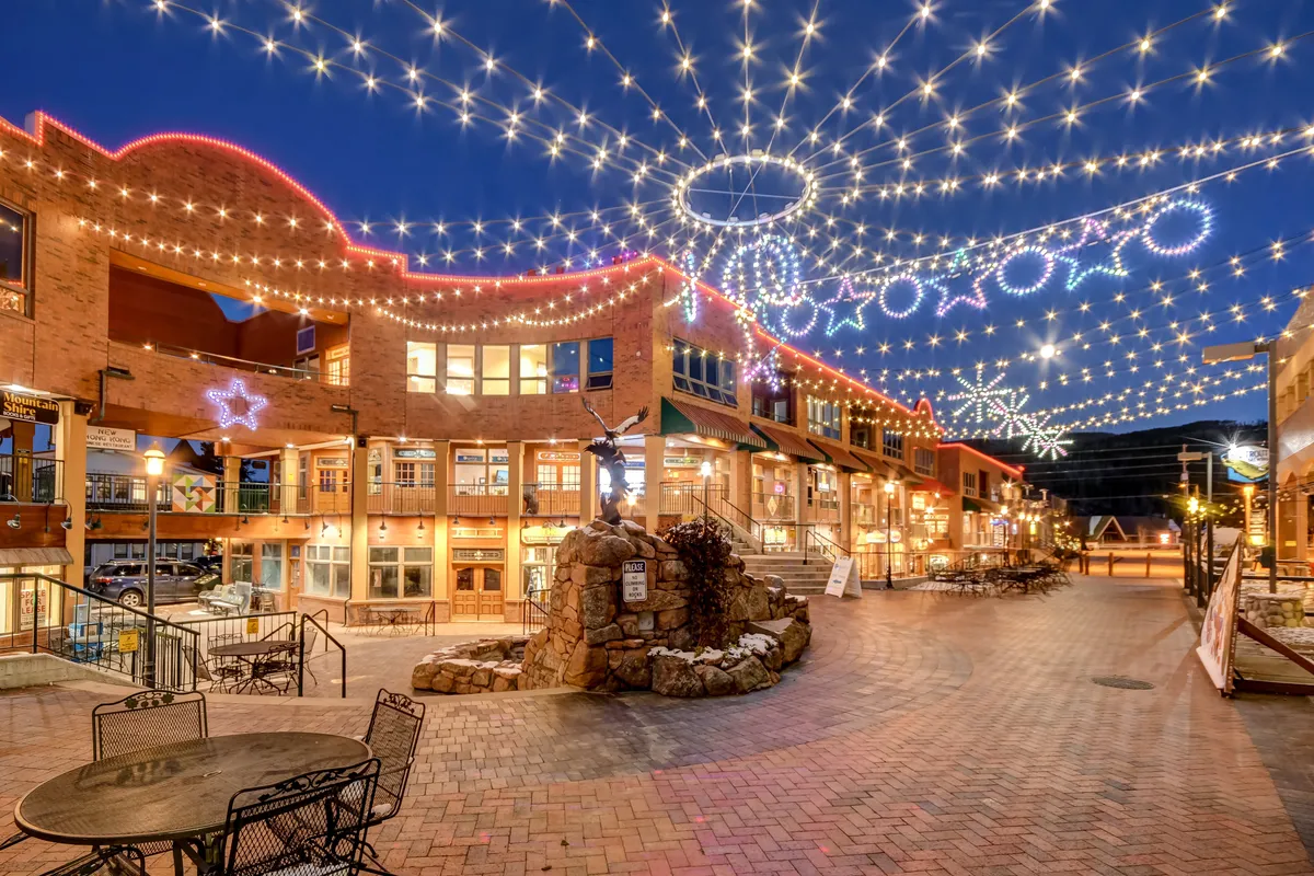 A lively plaza scene aglow with string lights and decorative stars, centered around a sculpture and surrounded by shops and restaurants. The area is set up for outdoor dining, shopping, and community gatherings, with a festive, active downtown vibe.