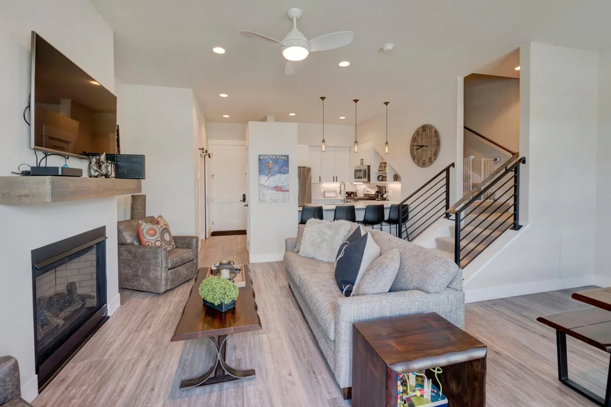 The photo offers a wide perspective on the central living space, featuring a fireplace, plush sofa, dining area, kitchen island, and a staircase. The warmth of the fire and soft lighting create a welcoming environment ideal for family gatherings and entertaining.