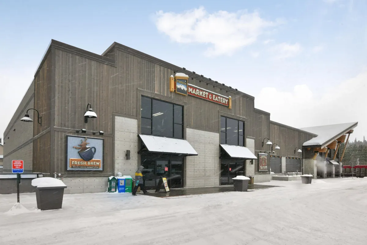 This image features the welcoming exterior of a market storefront, with clear signage, large windows, and an inviting entrance awning. The scene exudes a warm, communal atmosphere, suggesting convenience and accessibility for shopping or gathering, with hints of seasonal decorations and a snowy day outside.
