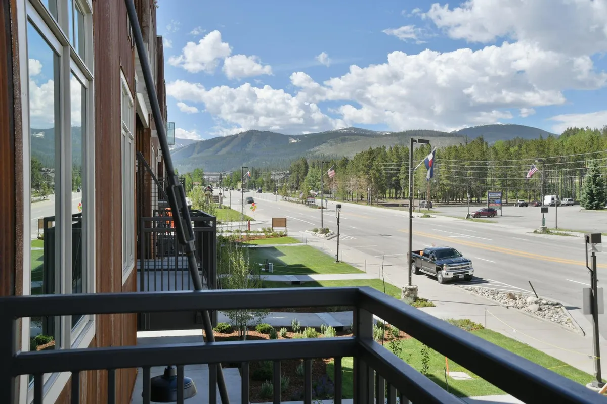 A private balcony with a railing overlooks a scenic mountain range, with a road and well-maintained landscaping visible below. The scene suggests tranquility and the perfect vantage point to soak in mountain views or watch sunsets.