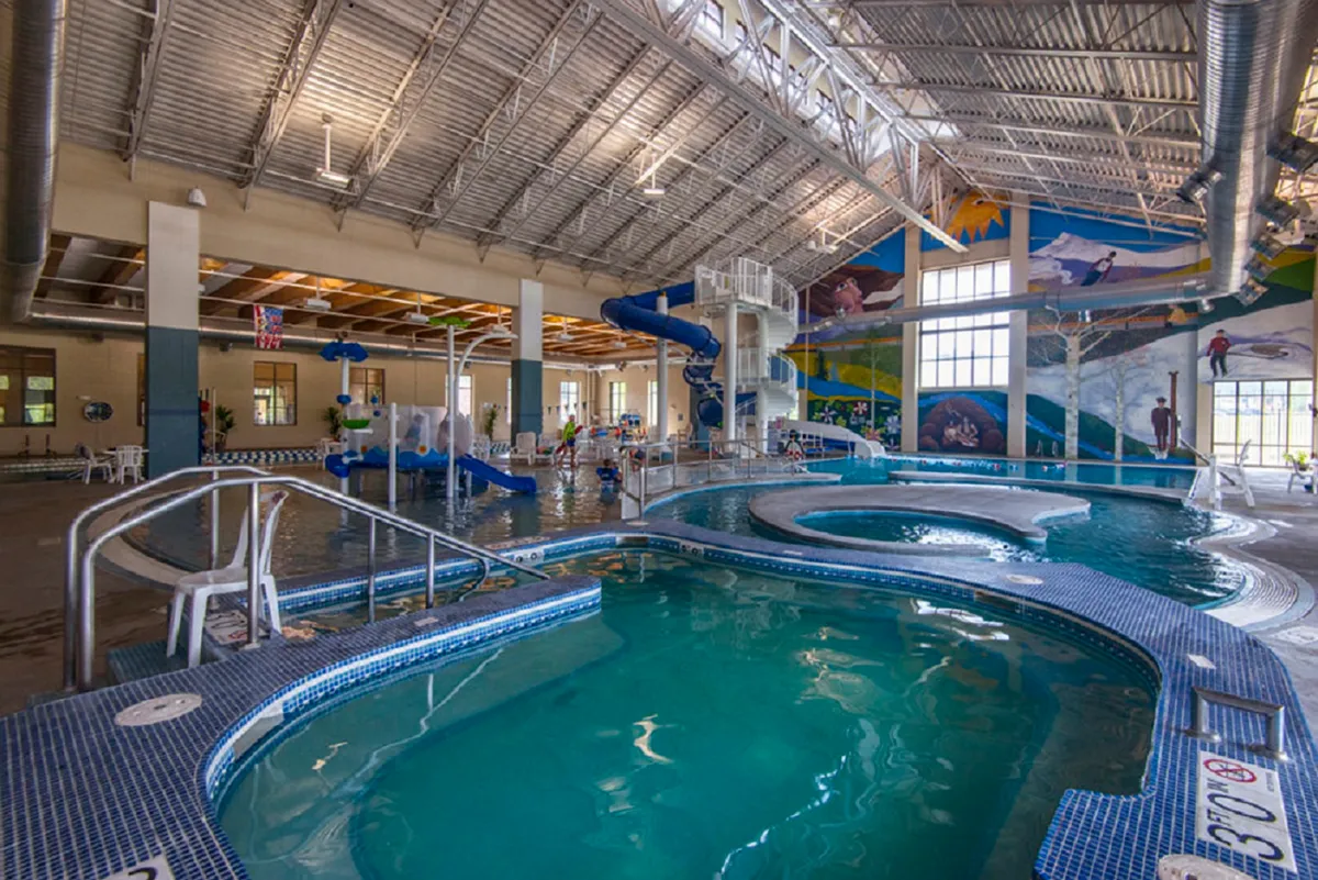 This image features a vibrant indoor pool area with a water slide, children playing, and a colorful mural on the wall. The atmosphere is energetic and joyful, emphasizing fun family activities and recreation.