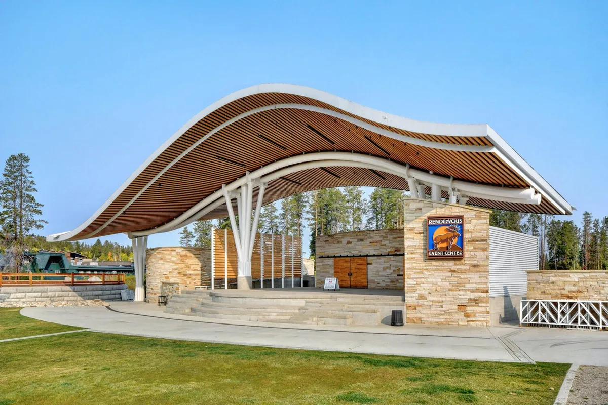 An exterior view of the Hideaway Park amphitheater, featuring a striking curved roof, stone facade, and wooden accents. The architecture is distinctive and the scene suggests a space designed for concerts, community events, and celebrations.