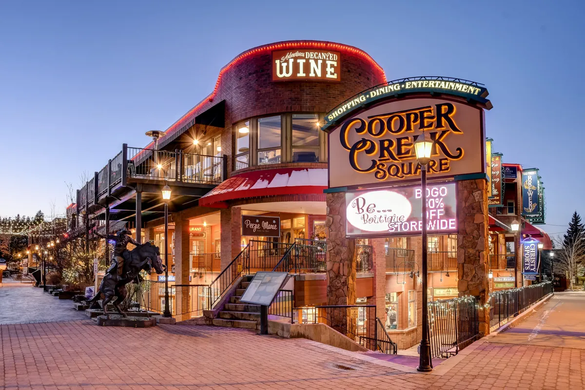 An exterior shot of Cooper Creek Square, featuring its prominent sign, a wine shop, and decorative lighting. The scene exudes a festive, communal mood and hints at local shopping, dining, and entertainment options available nearby.