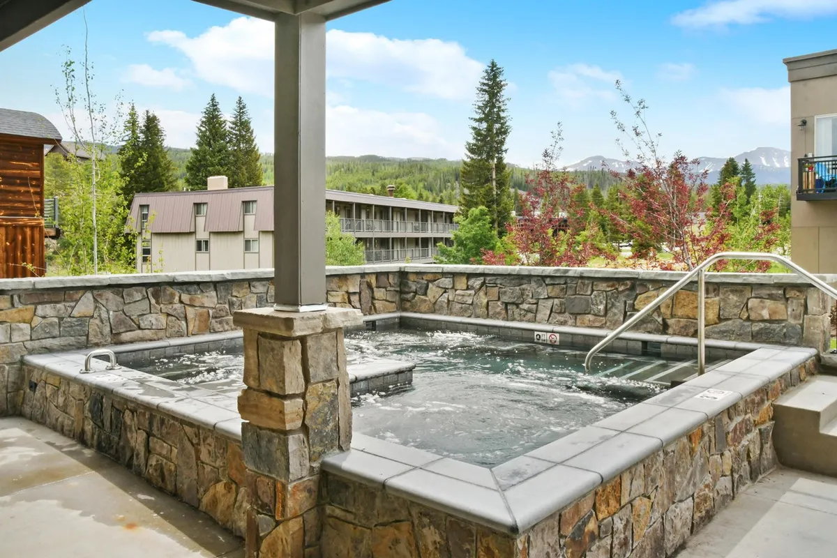 A large, inviting hot tub set outdoors with a stunning mountain backdrop. The area is surrounded by stonework and natural landscape, conveying a sense of relaxation and rejuvenation, ideal for socializing or winding down after outdoor activities.
