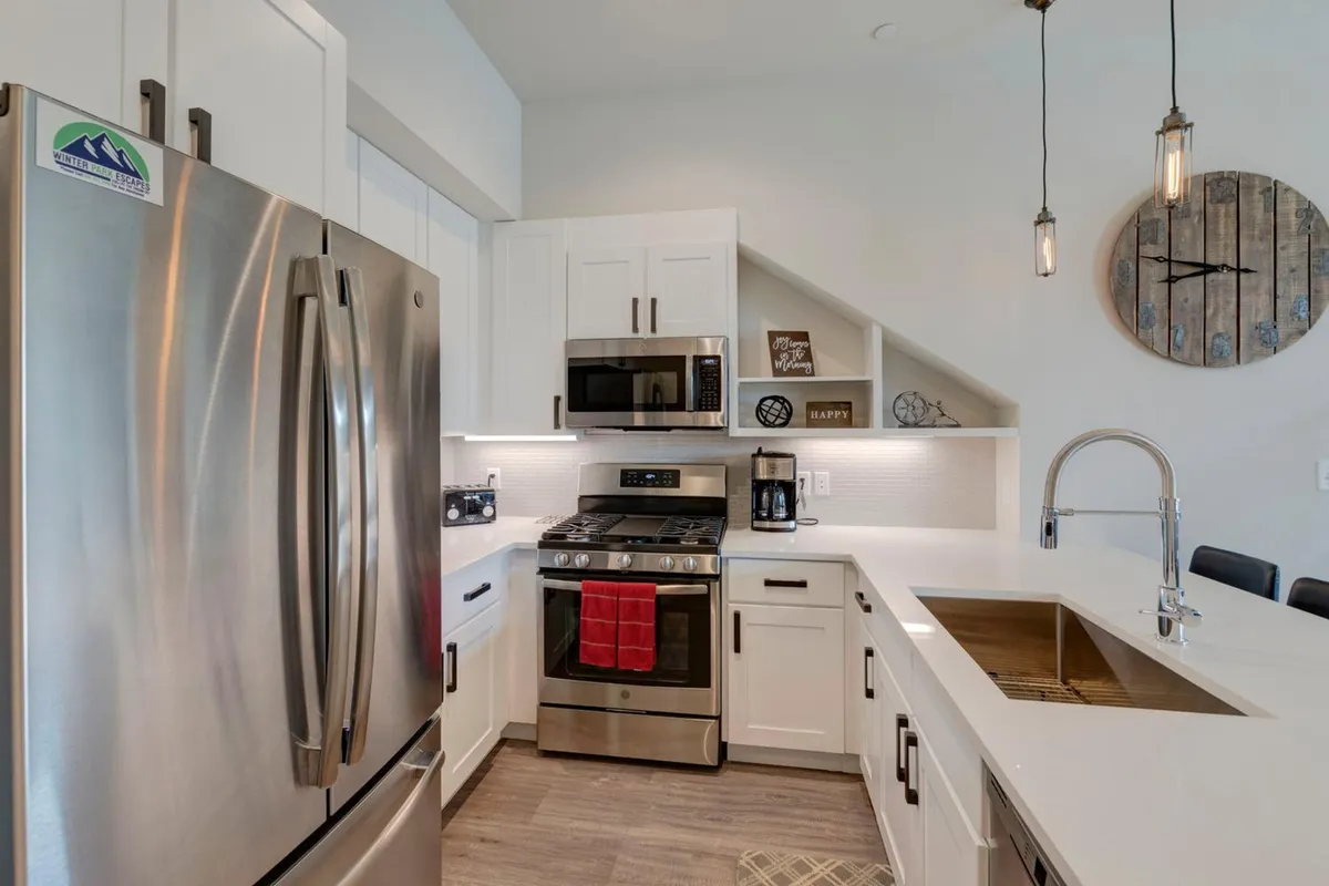 A spacious, modern kitchen featuring a stainless steel refrigerator, sleek stove and oven, minimalist kitchen island, and wooden clock on the wall. Decorative wall art and subtle accents add warmth, while the open layout invites both cooking and socializing.